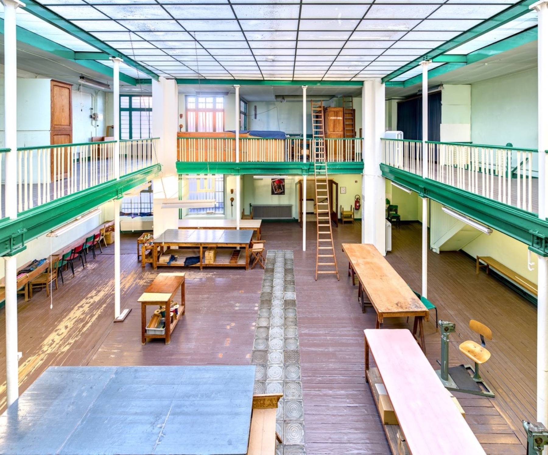 De grote hal van de Paraplufabrique vandaag, met werktafels, een houten ladder en een galerij met groene balken